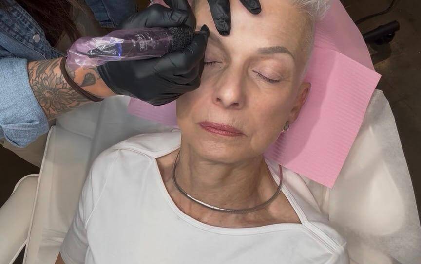 Breast Cancer Sucks. 6 Surprising Ways to Support Survivors - 4 A person with short gray hair lies with eyes closed as a technician in gloves uses a tattoo device near their eyebrow, a procedure that can be especially meaningful for breast cancer survivors seeking to restore confidence and a sense of normalcy.