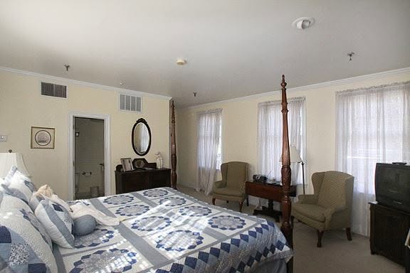 A bedroom in one of the South’s most haunted hotels, featuring a four-poster bed, two armchairs, a dresser with a mirror, and a TV on a stand. An open door leads to the bathroom while light streams in through sheer curtains, hinting at secrets whispered by lingering spirits.