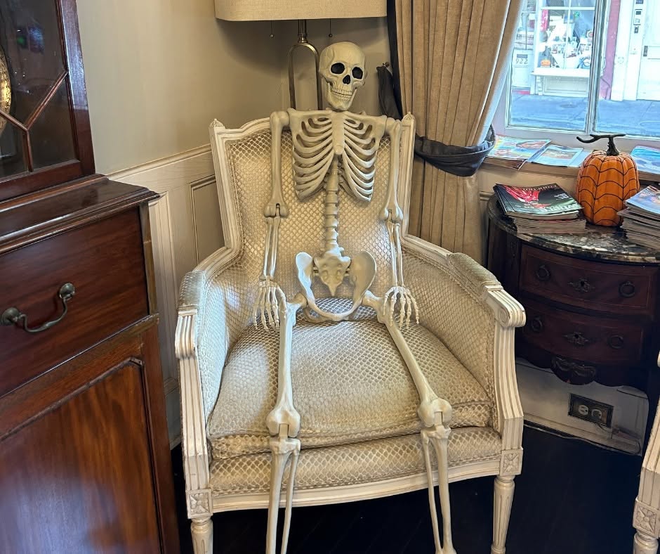 A human skeleton prop is seated in an upholstered armchair in a corner of a room decorated for Halloween, evoking the mysterious ambiance often found in the most haunted hotels of the South.