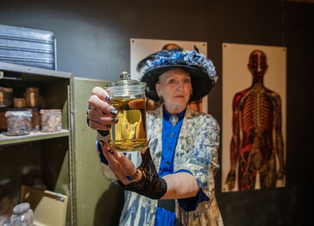 A woman in vintage clothing holds a glass jar of yellow liquid in a room with anatomical posters and shelves of jars - one of the most haunted hotels in the South.