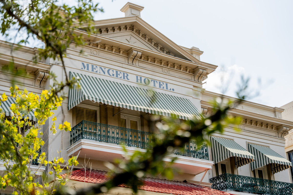 Facade of Menger Hotel with green-and-white striped awnings, balconies, and leafy branches partially in the foreground—an iconic sight in the South, and widely regarded as one of its most haunted hotels.