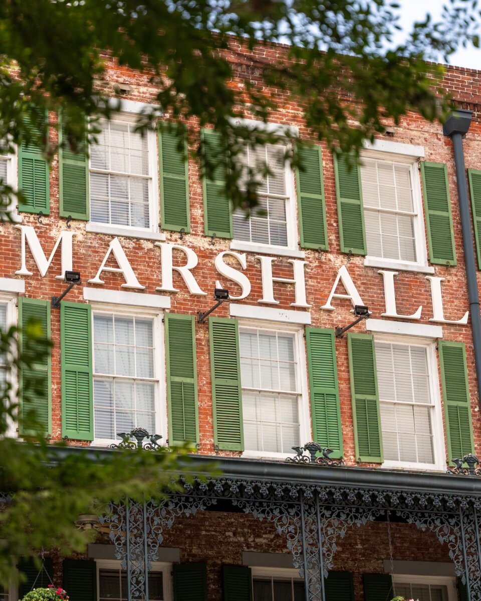 Brick building with green shutters and large white letters spelling “MARSHALL” on the facade, partially obscured by tree branches—this historic site is renowned as one of the most haunted hotels in the South.