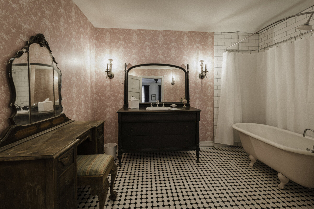 Vintage-style bathroom with pink wallpaper, black vanity, wooden dressing table, clawfoot bathtub, and black-and-white tile floor.