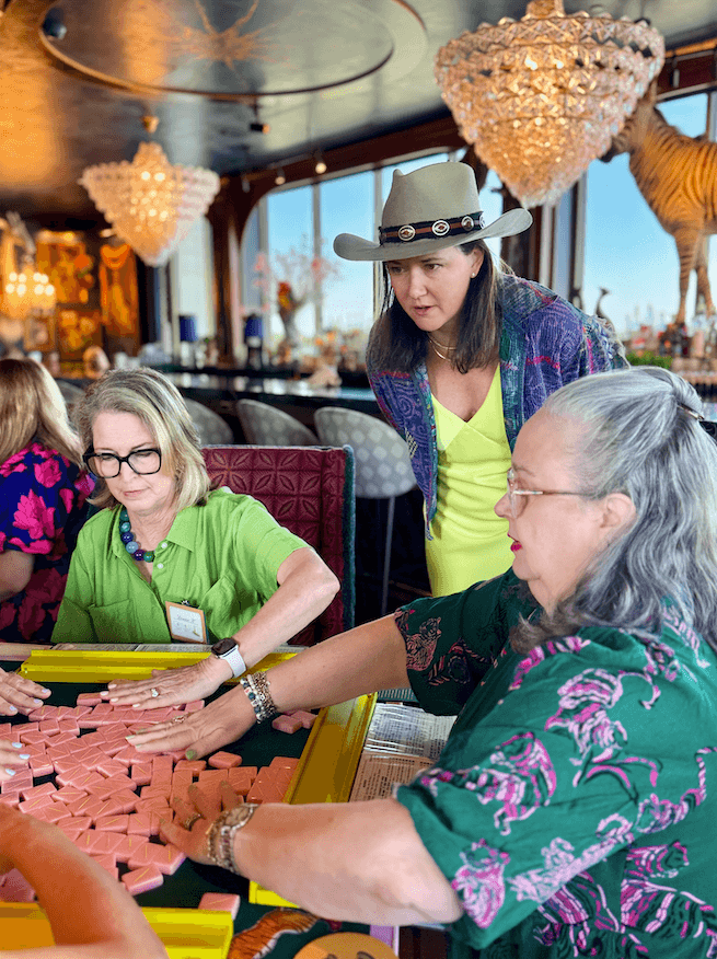 Your Ultimate Memphis Mahjong Guide - 2 Three women gather around a table, arranging pink game tiles as they consult a Memphis Mahjong guide. One woman stands and observes while the other two are seated, focused on the game. Chandelier lights and decor fill the background.