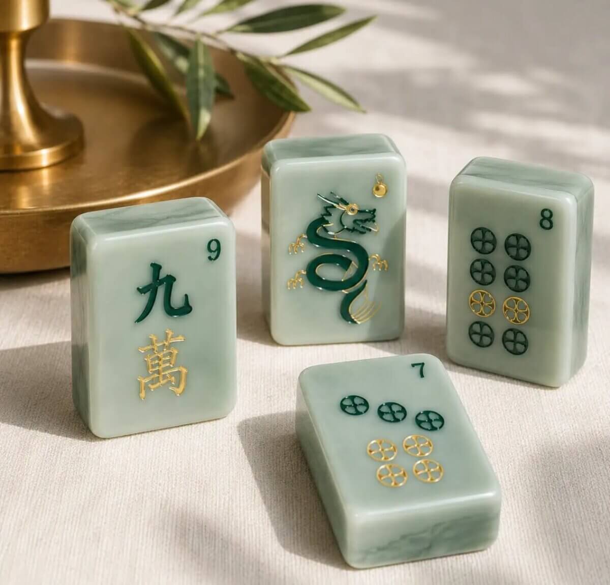 Your Ultimate Memphis Mahjong Guide - 7 Four jade Mahjong tiles, including a green dragon and the numbers 7, 8, and 9, are arranged on a light surface beside a brass tray and olive branch.
