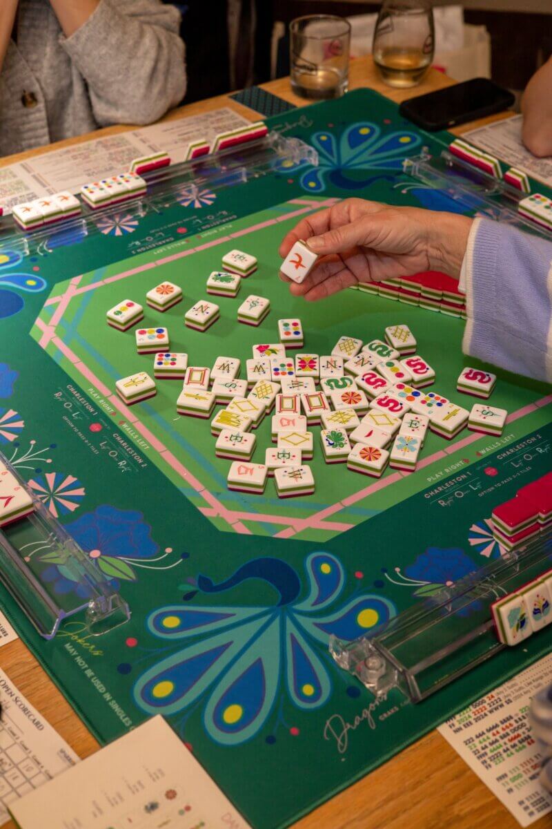 Your Ultimate Memphis Mahjong Guide - 4 Four people play Mahjong around a green table, with tiles scattered and scorecards visible. One person is picking up a tile from the center. Drinks and a phone are on the table—an inviting scene that could easily be part of any Memphis Mahjong gathering.