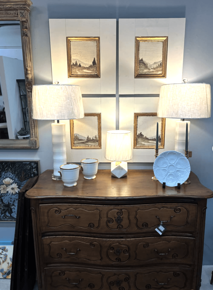 Your Guide to Antiquing in Memphis: 10 Shops Worth Exploring - 5 A wooden dresser with ornate carving holds two white lamps, a small central lamp, three cups, and a white plate; framed art hangs on the wall above.
