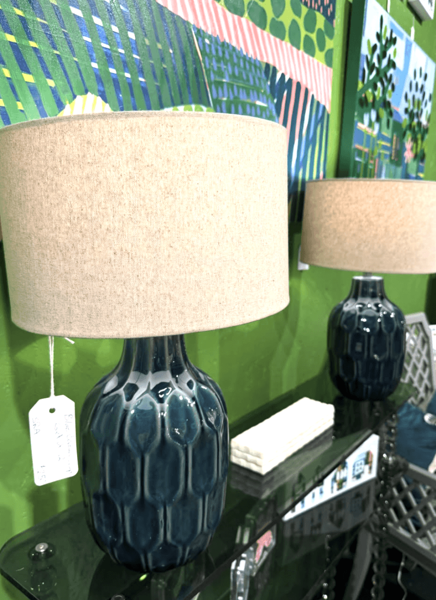 Your Guide to Antiquing in Memphis: 10 Shops Worth Exploring - 11 Two table lamps with textured dark blue ceramic bases and beige fabric lampshades sit on a glass shelf against a green wall decorated with colorful paintings.