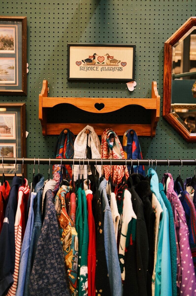Your Guide to Antiquing in Memphis: 10 Shops Worth Exploring - 10 A rack of colorful vintage clothes hangs below a wooden shelf and framed “Rejoice Always” cross-stitch on a green pegboard wall, with framed art and a mirror on either side.