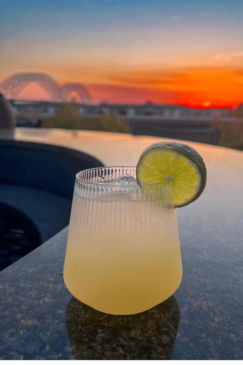 Did You Know You Could Spend the Night in the Memphis Pyramid? - 2 A glass of yellow cocktail with ice and a lime slice sits on a granite surface at sunset, with a bridge and vibrant sky in the background.