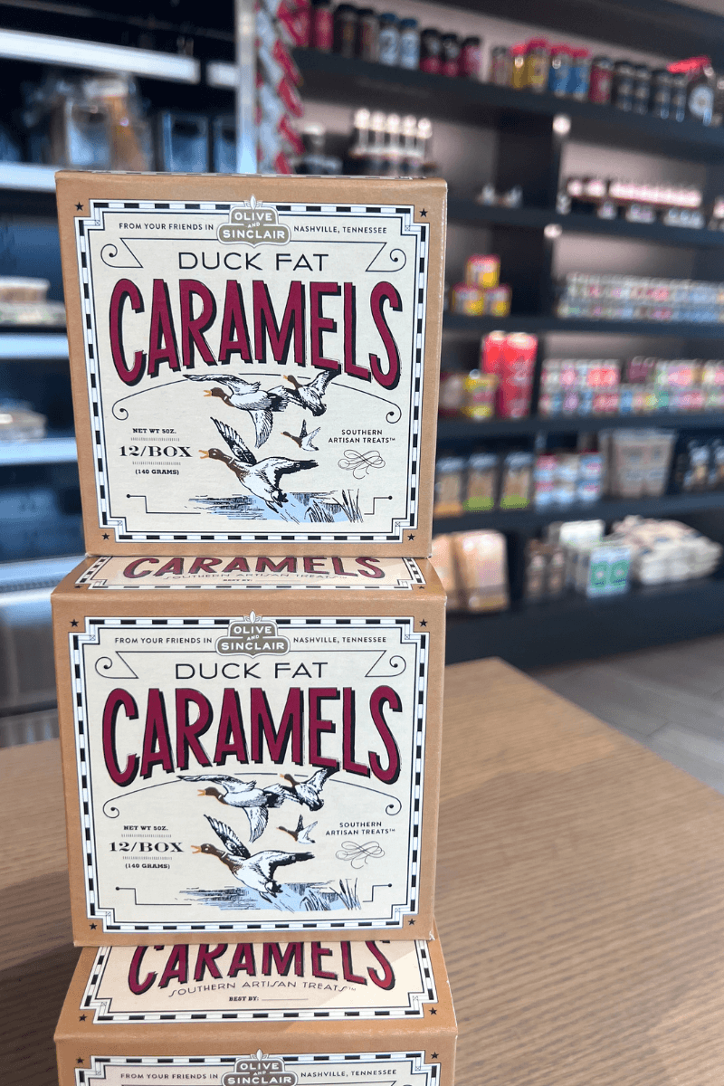 Three boxes of Olive & Sinclair Duck Fat Caramels are stacked on a wooden table in a specialty food store. Shelves with various products are visible in the background.