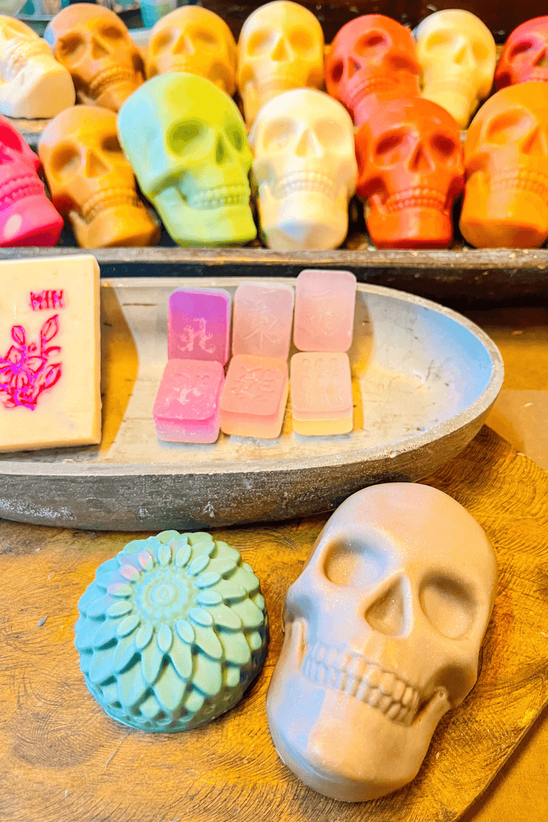 Colorful skull-shaped soaps, pastel rectangular soaps, a floral round soap, and a beige skull soap are displayed on wooden trays.