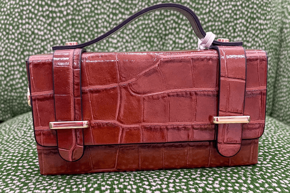 A rectangular, brown, crocodile-embossed leather handbag with a black handle and gold-tone hardware sits on a green and white patterned surface.