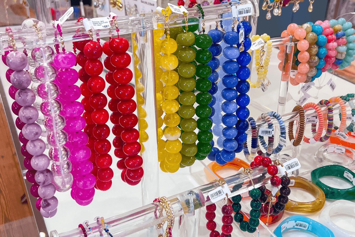 Colorful bead bracelets are displayed on a clear acrylic rack, organized by color groups including pink, red, yellow, green, blue, and purple. Several round bangle bracelets are below.