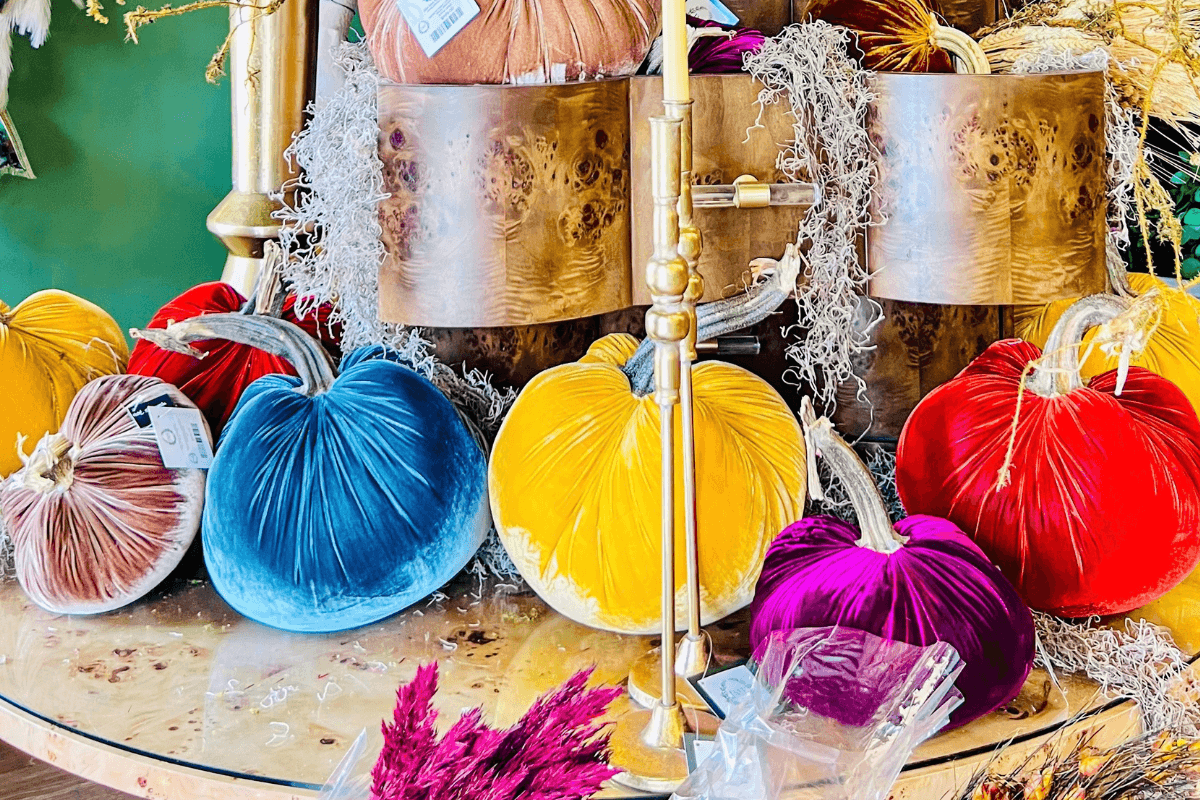 Plush velvet pumpkins in various bright colors are arranged on a glass shelf with decorative moss and wooden containers in the background.