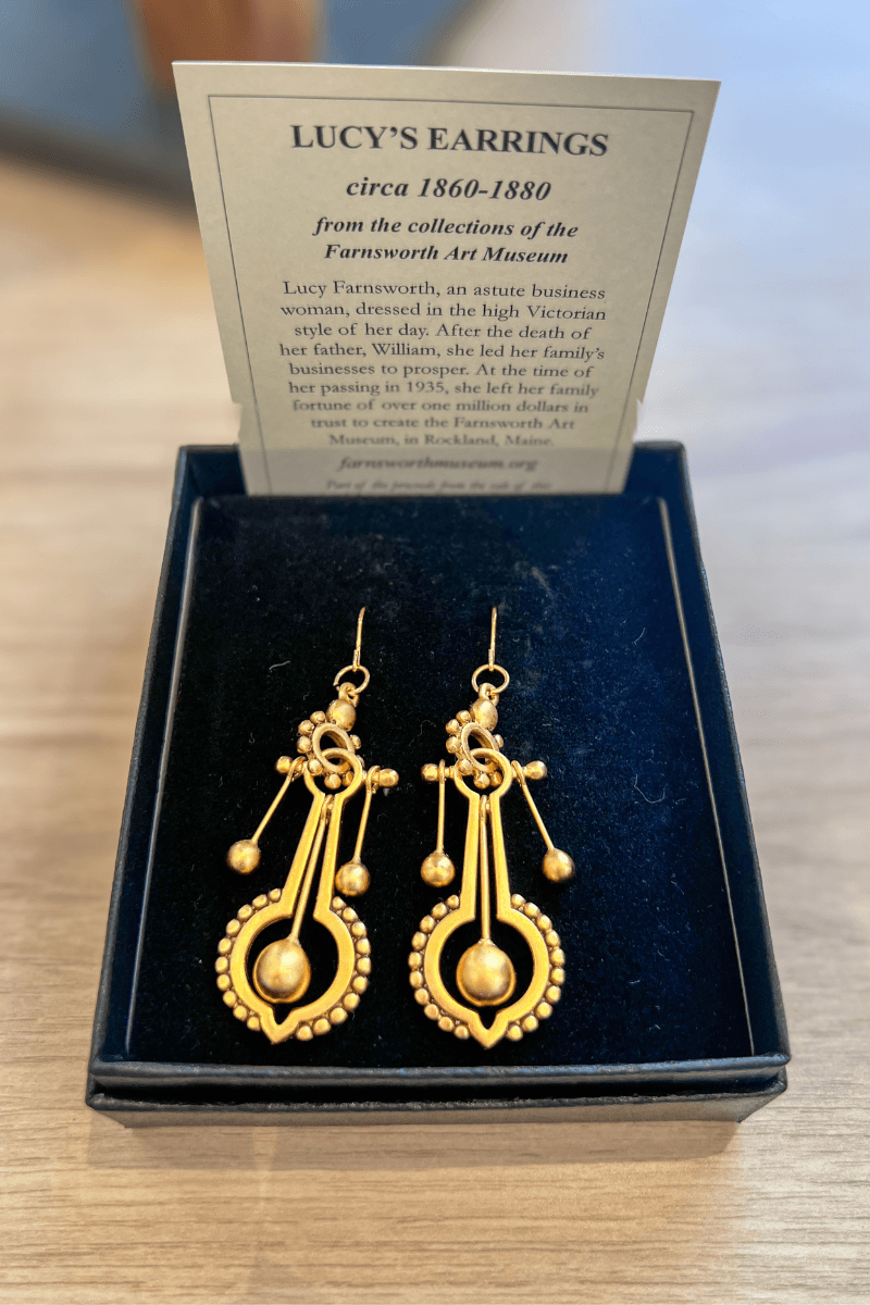A pair of ornate gold earrings in a black box, with a museum information card about “Lucy’s Earrings” from circa 1860–1880 displayed behind them.