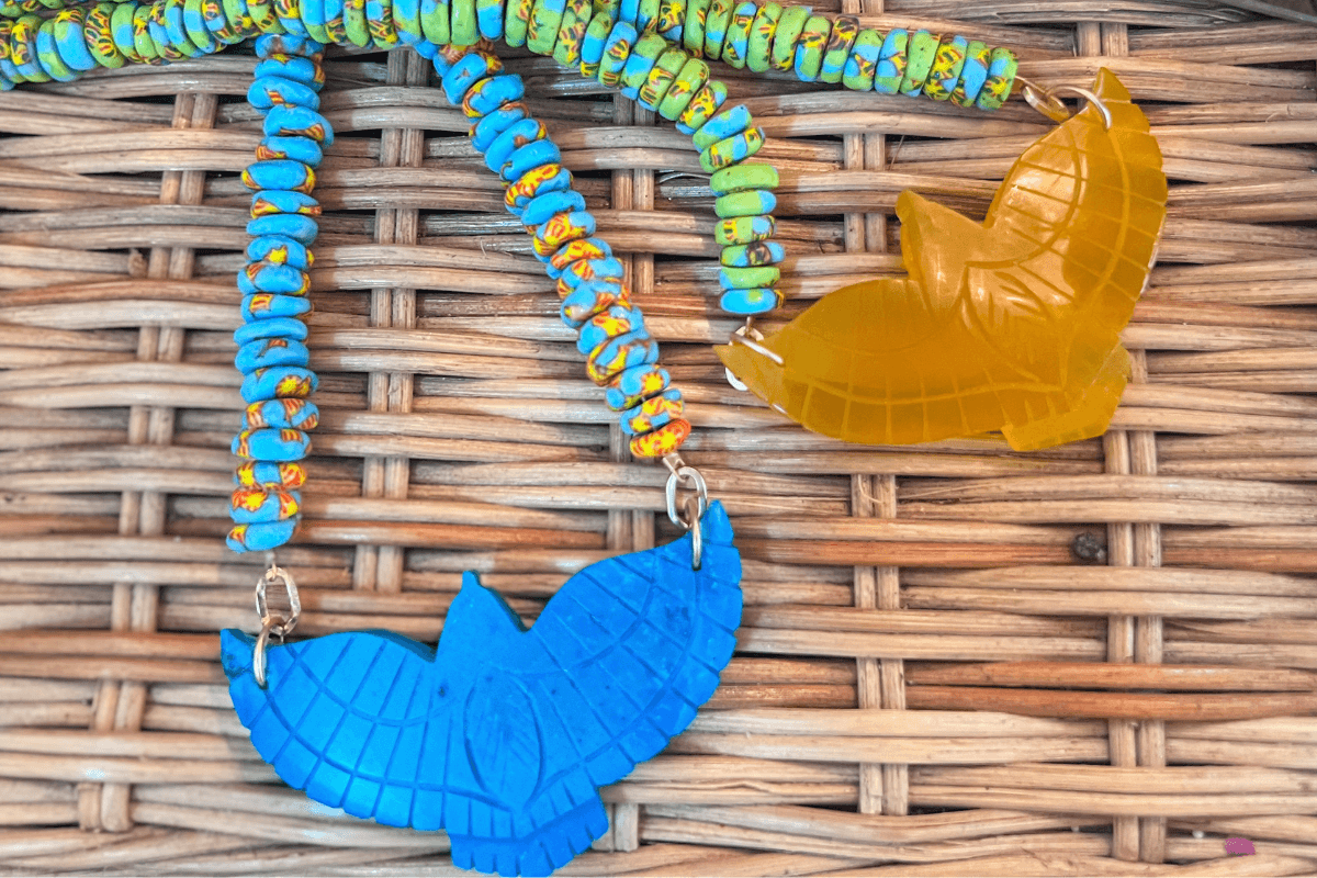 Two necklaces with bird-shaped pendants, one in blue and one in yellow, rest on a woven wicker surface. The blue necklace features turquoise and yellow beads.