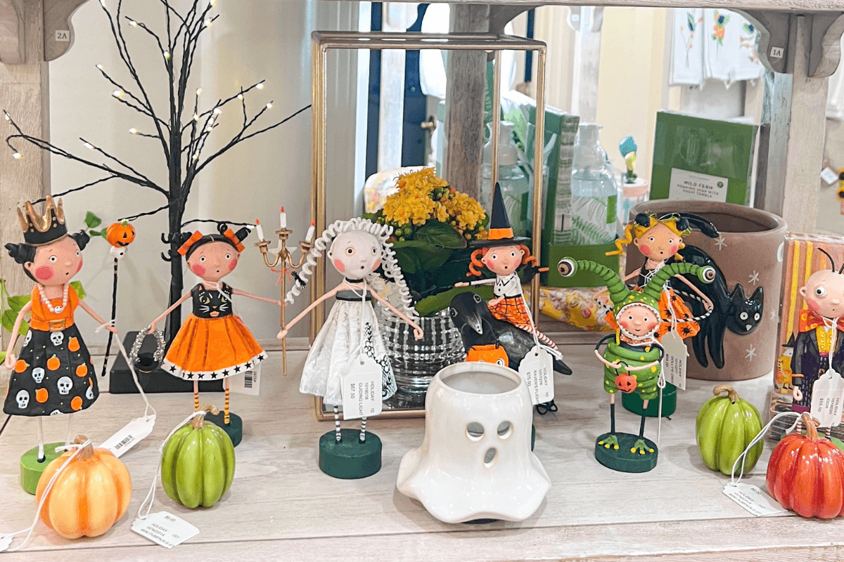Five Halloween-themed figurines and a white ghost candle holder are displayed on a wooden shelf, surrounded by small pumpkin decorations and a decorative tree with lights.