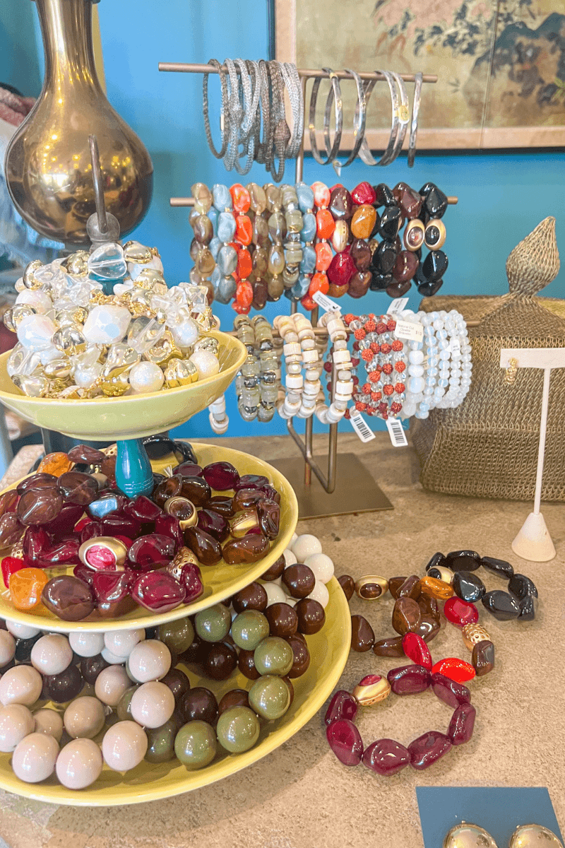 Assorted colorful stone bead bracelets and bangles displayed on a tiered stand and metal rack on a beige countertop in a boutique.