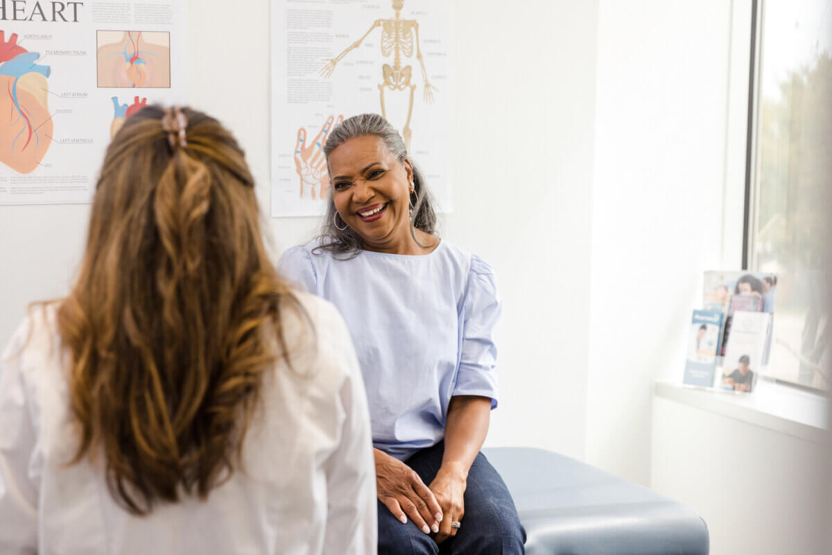 Your Ears May Itch — and Other Surprising Things About Menopause - 3 Smiling patient sits on an examination table, discussing menopause facts with a healthcare professional. Medical posters and pamphlets are visible in the background.