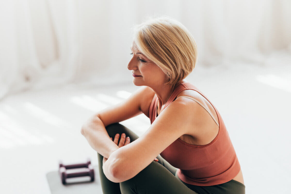 Your Ears May Itch — and Other Surprising Things About Menopause - 4 A woman with short blonde hair in workout clothes sits on the floor with arms around her knees, reflecting on perimenopause. A pair of dumbbells is placed nearby.