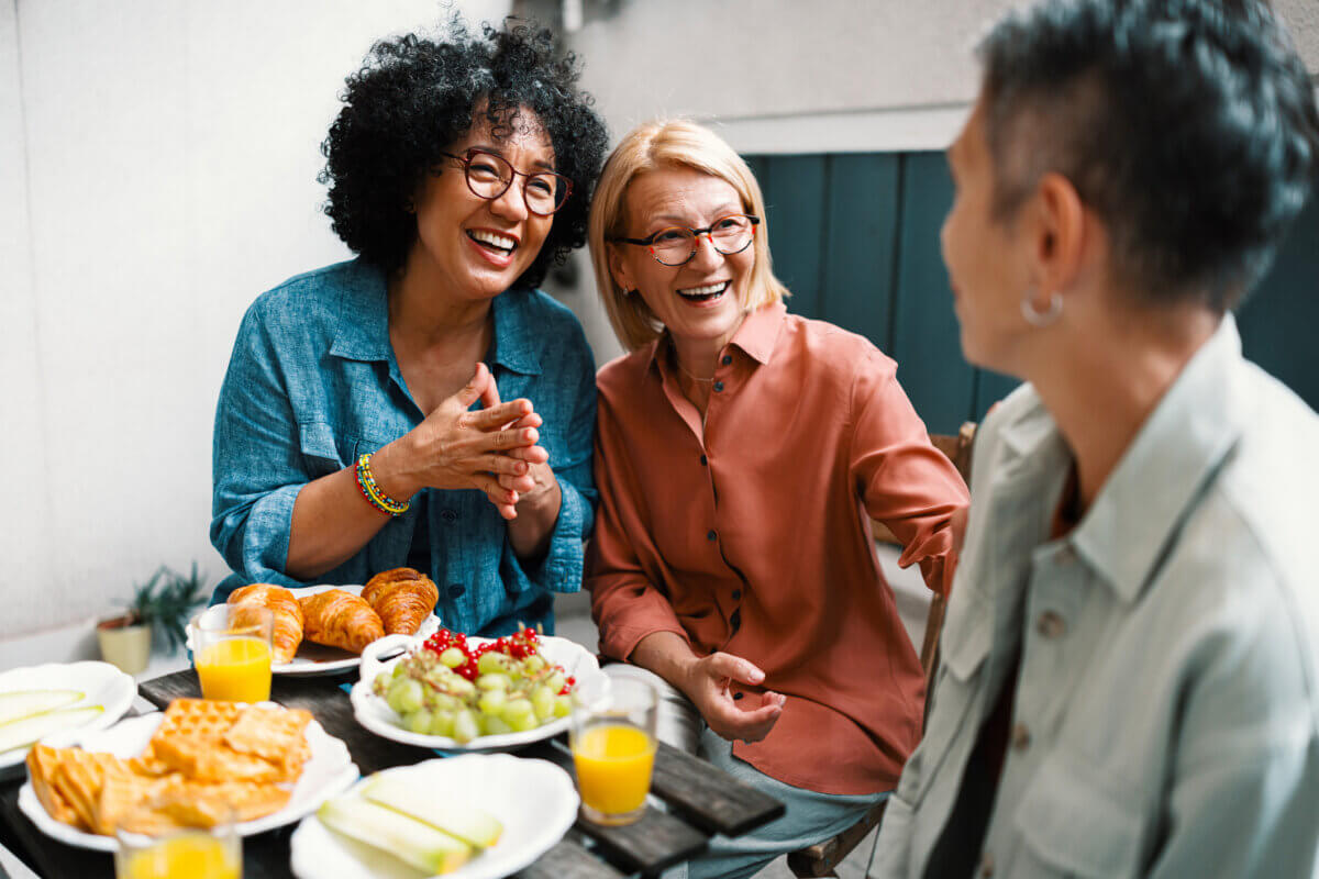 Your Ears May Itch — and Other Surprising Things About Menopause - 2 Three older adults sit at an outdoor table with breakfast foods and orange juice, smiling and sharing surprising facts about menopause together.