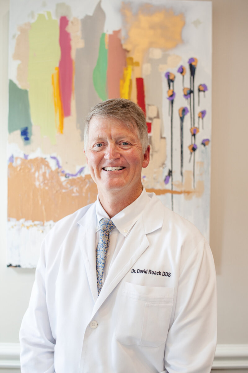 Healthy Smiles for Life: A Guide to Family Dental Care For Every Age - 2 A man in a white lab coat with “Dr. David Roach DDS” embroidered stands smiling in front of an abstract painting, embodying the commitment to family dental care and healthy smiles.