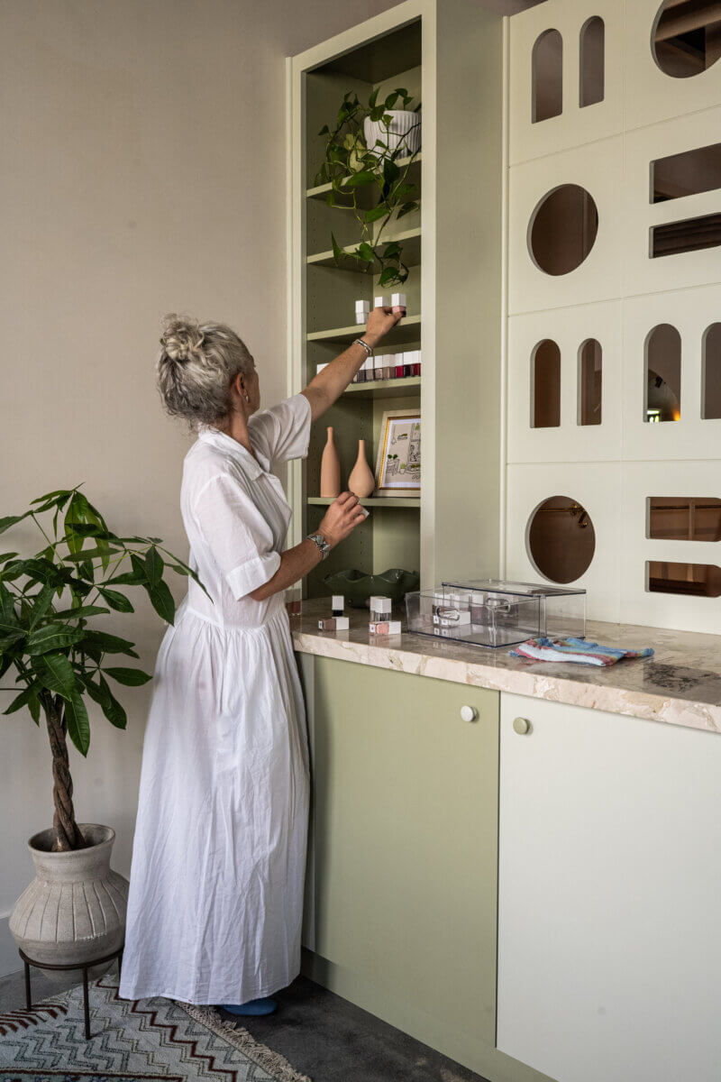Person in a white dress arranges a plant on a tall green shelf beside a countertop with small boxes, vases, and a potted plant on the floor.