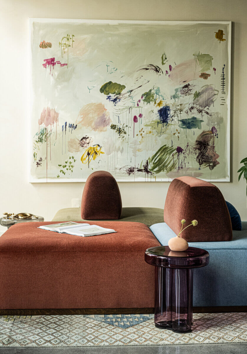 Modern living room with a red and blue sectional sofa, a round purple side table, an open book, and an abstract painting on the wall.