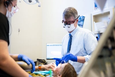 A dentist wearing magnifying glasses and gloves performs a dental procedure on a patient, assisted by another person, in a brightly lit clinic dedicated to family dental care and healthy smiles.