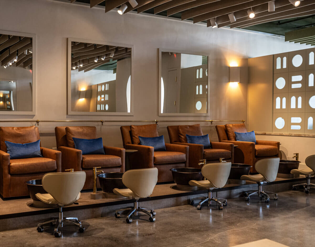 A row of brown leather chairs with blue cushions faces manicure stations with beige stools in a modern, well-lit salon.