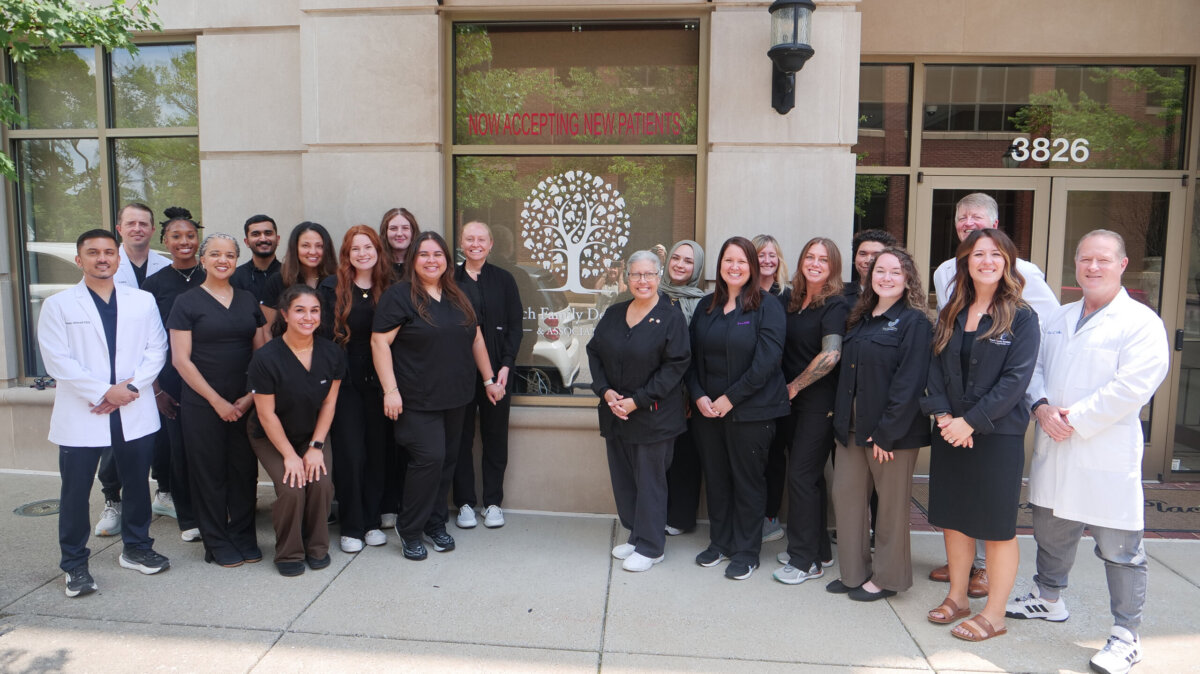 Healthy Smiles for Life: A Guide to Family Dental Care For Every Age - 6 A group of dental staff in black uniforms and white coats stand smiling outside a clinic entrance with a tree logo and a “Now Accepting New Patients” sign, ready to welcome you and your loved ones with expert family dental care for healthy smiles.