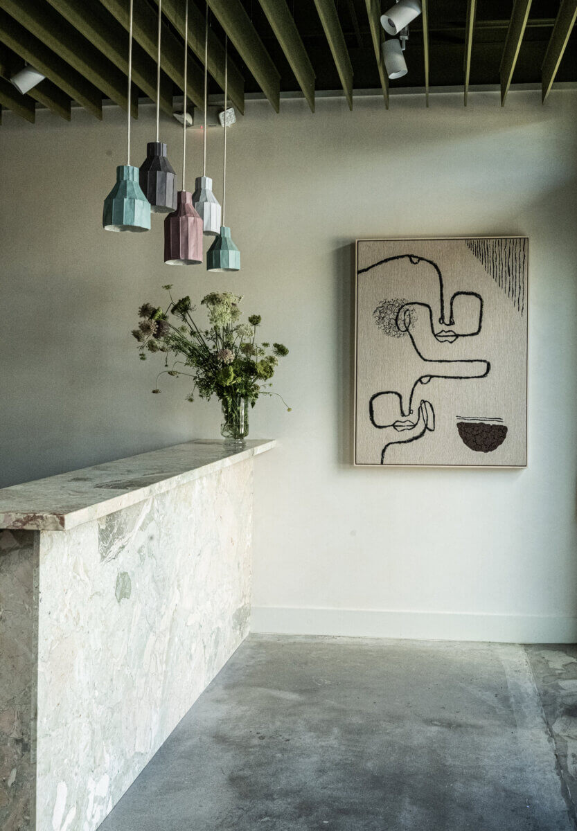 Modern interior with a marble counter, hanging geometric pendant lights, a vase of flowers, and abstract line art on a white wall.