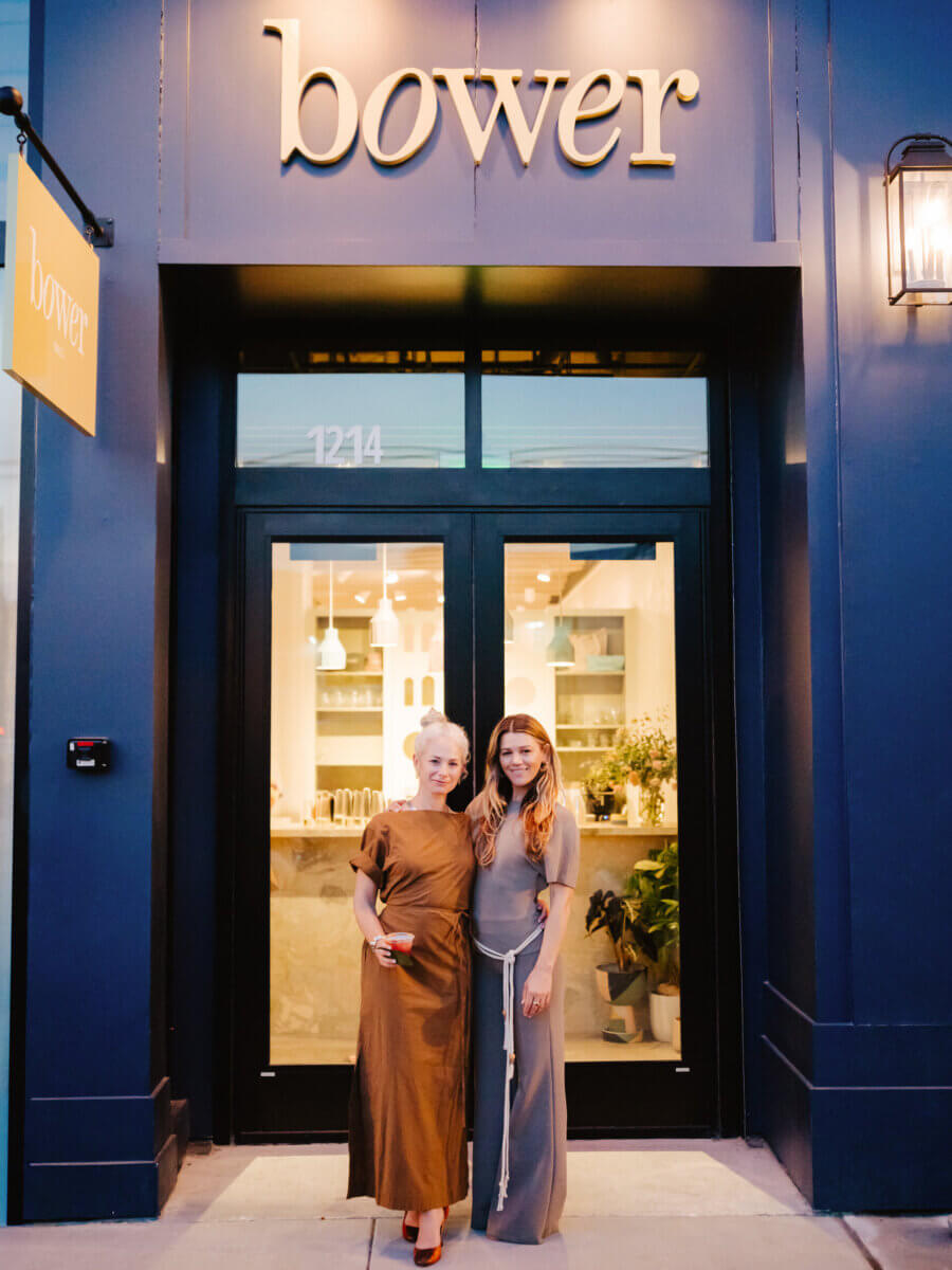 Two women stand side by side, smiling in front of a glass door entrance to a building with a “bower” sign above them.