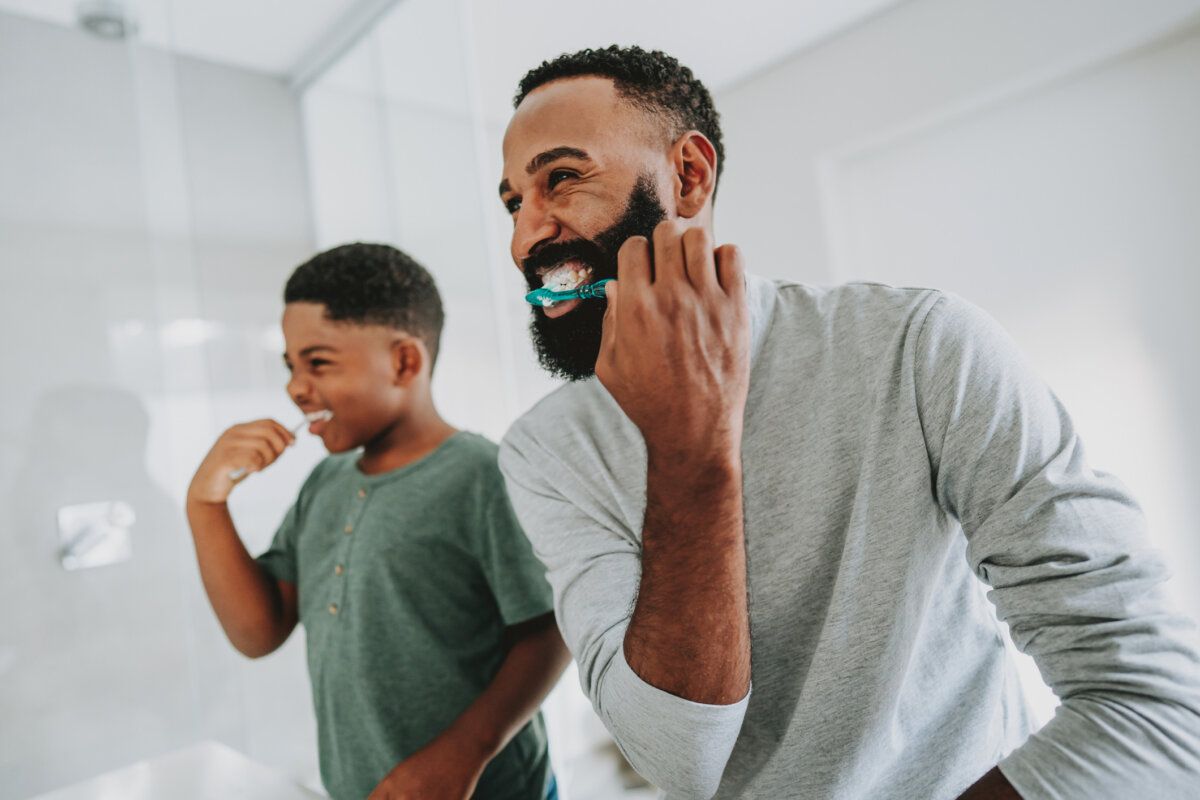 Healthy Smiles for Life: A Guide to Family Dental Care For Every Age - 3 An adult and a child stand in front of a bathroom mirror, brushing their teeth and smiling, enjoying a moment of family dental care for healthy smiles.
