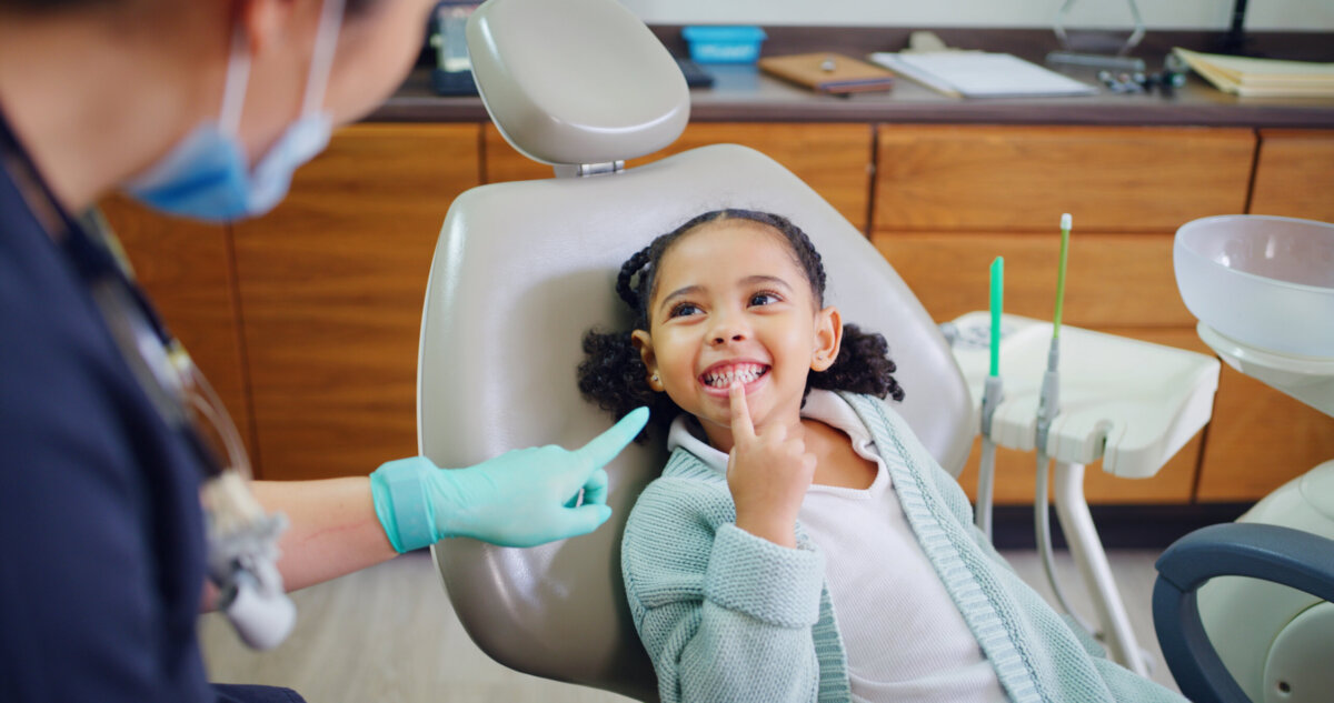 Healthy Smiles for Life: A Guide to Family Dental Care For Every Age - 4 A young child sits in a dental chair smiling and pointing at their teeth while a dental professional wearing gloves interacts with them, highlighting the importance of healthy smiles as part of family dental care.