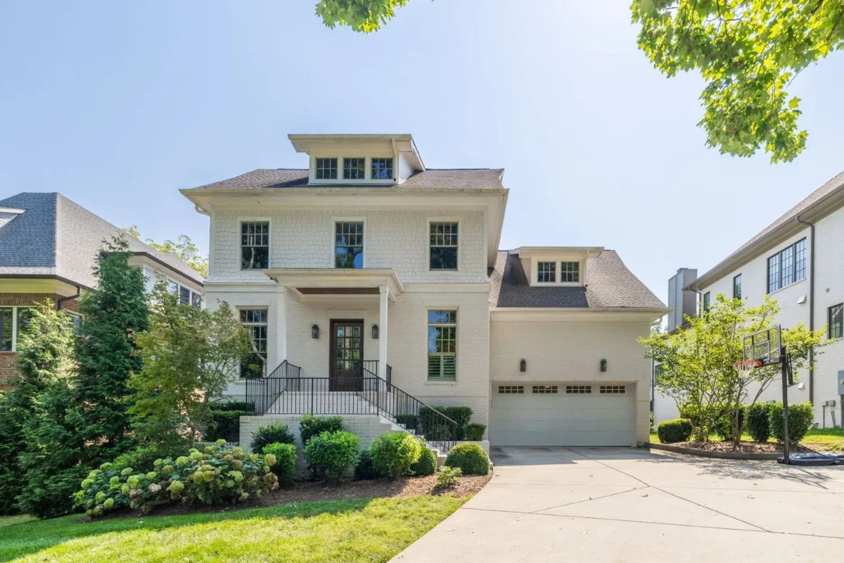 Elegant two-story white brick house with black trim, a welcoming front porch, and an attached two-car garage located at 4000 Wallace Lane, Nashville. The beautifully landscaped yard features a basketball hoop, all set under bright sunny skies.