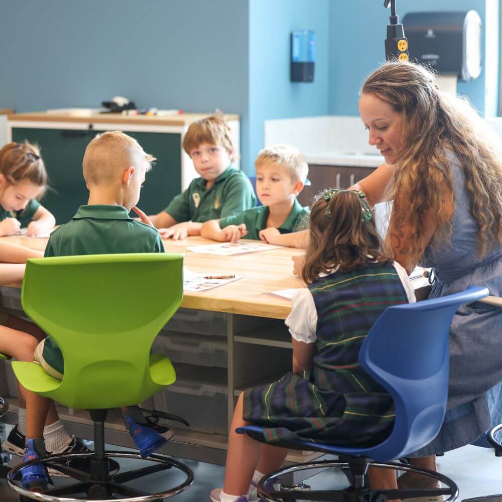 This Nashville School Is Giving Students More Room to Explore, Create & Build - 6 A teacher assists young students seated around a table at a Nashville school, engaged in classroom activities. The children, who may have learning differences, wear green uniforms and colorful chairs are visible.