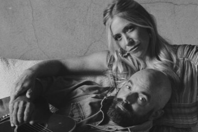 A woman with long blonde hair sits on a couch, resting her arm on the shoulder of a bearded man playing a guitar. Both are looking toward the camera in this black-and-white photo, capturing the essence of creative partnerships reminiscent of artists like Drew Holcomb and his collaborations.
