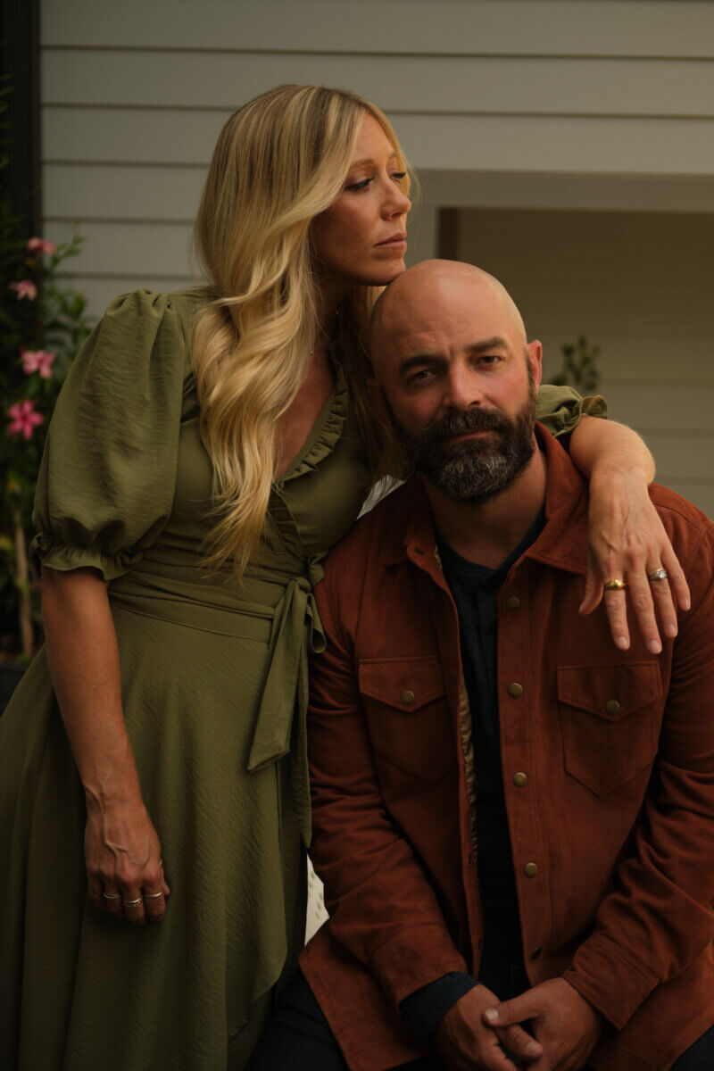 A woman in a green dress stands with her arm around a seated man in a brown jacket in front of a house, both looking serious, capturing the essence of creative partnerships reminiscent of Drew Holcomb singing with his spouse.