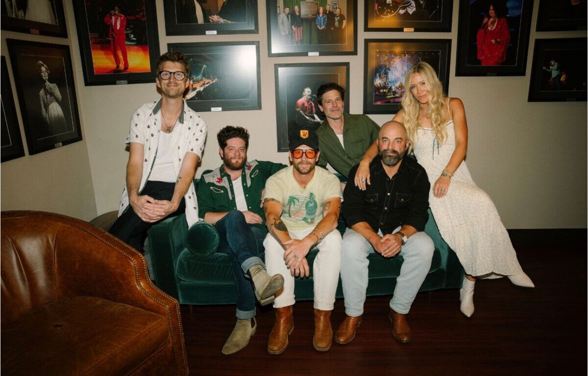 Six people pose together on and around a green couch in front of framed concert photos on a wall.