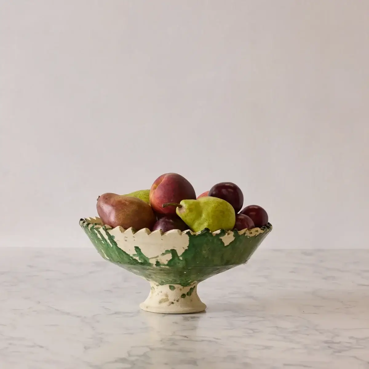 A green and white ceramic pedestal bowl filled with pears, apples, and plums sits on a marble surface against a plain light background—a charming September FINDS from Nashville perfect for adding seasonal elegance to your home.