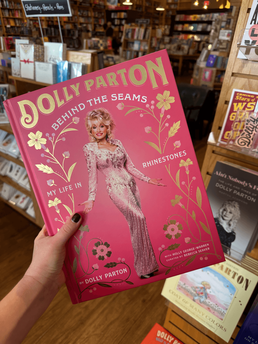 A hand holds the book “Dolly Parton: Behind the Seams – My Life in Rhinestones” in a Nashville bookstore, with shelves of books visible in the background.