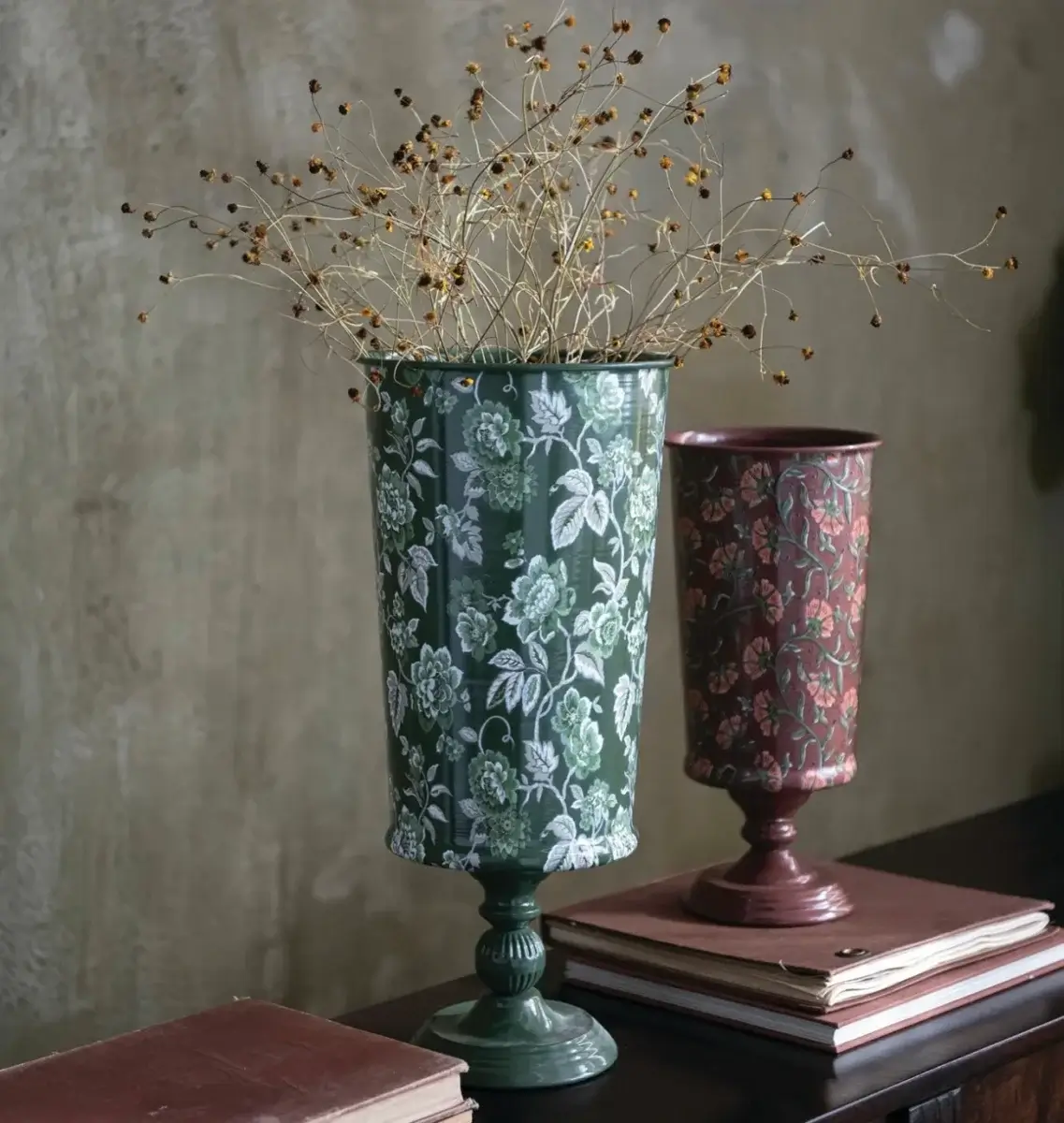 Two patterned metal vases with dried stems inside, placed on a dark surface with stacked books—these September FINDS bring a touch of Nashville charm to any space, set beautifully against a textured wall.