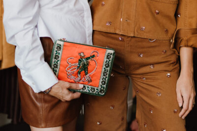 Two people stand close together; one holds a colorful clutch with a cowboy design, reminiscent of unique finds from Nashville boutiques. Both wear brown or tan clothing with decorative details.
