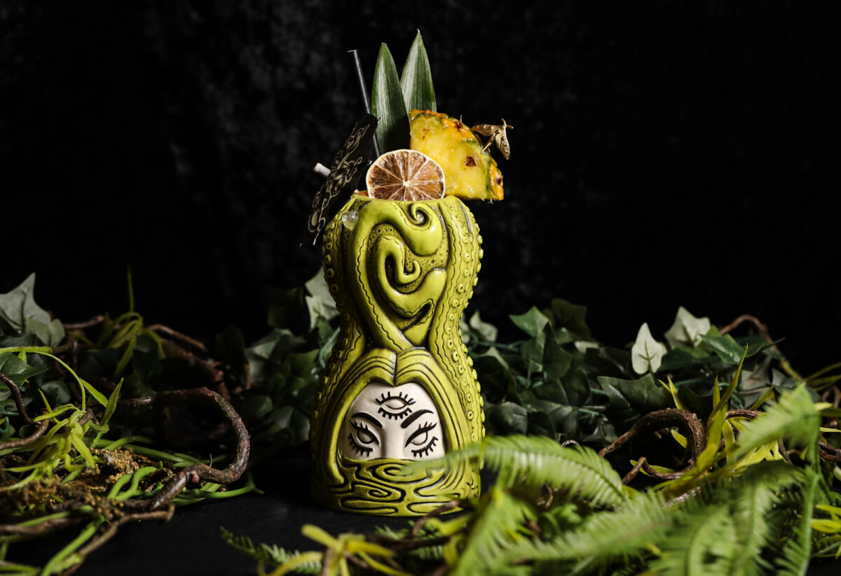 Green tiki cocktail in an ornate mug with an illustrated face, garnished with pineapple, dried lime, and leaves, surrounded by green foliage against a black background—a perfect fit for Nashville’s pop-up bars or anyone seeking a drink with dramatic flair.