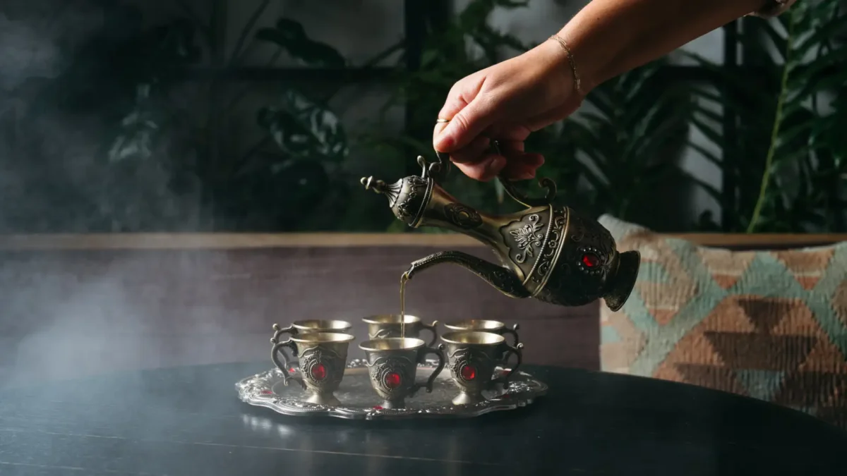 A hand pours tea from an ornate metal teapot into small decorated cups on a tray, with steam rising in the background—a cozy scene reminiscent of Nashville’s seasonal bars and their warm, inviting atmospheres.