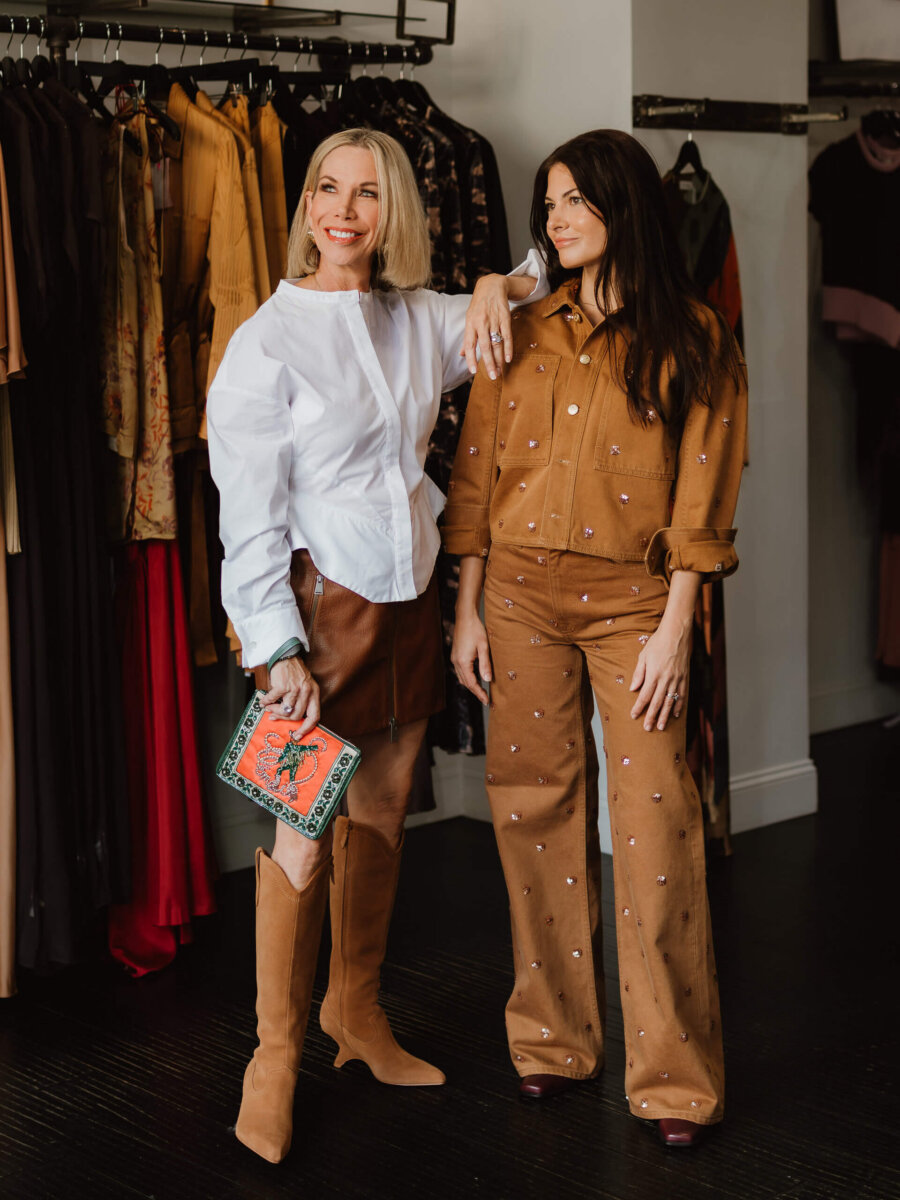 We’re Obsessed With This Rhinestone Cowboy Clutch (and 5 Other Nashville Fall Styles) - 2 Two women stand in a clothing store; one wears a white blouse with a brown skirt and boots, holding a clutch, while the other dons a brown pantsuit with embellishments. Dresses hang behind them.