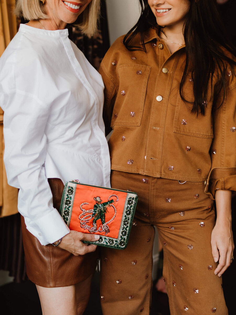 We’re Obsessed With This Rhinestone Cowboy Clutch (and 5 Other Nashville Fall Styles) - 3 Two women stand side by side; one wears a white blouse and brown skirt, the other a tan outfit—both showcasing styles found in Nashville boutiques. One holds a red and green embellished clutch bag. Only the lower faces and torsos are visible.