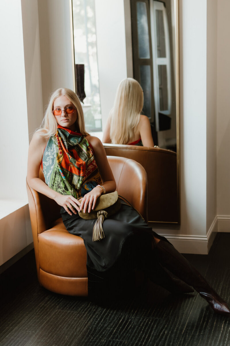 We’re Obsessed With This Rhinestone Cowboy Clutch (and 5 Other Nashville Fall Styles) - 4 Woman in a colorful scarf and sunglasses, styled with flair, sits on a brown chair by a window; her reflection is visible in a tall mirror behind her.